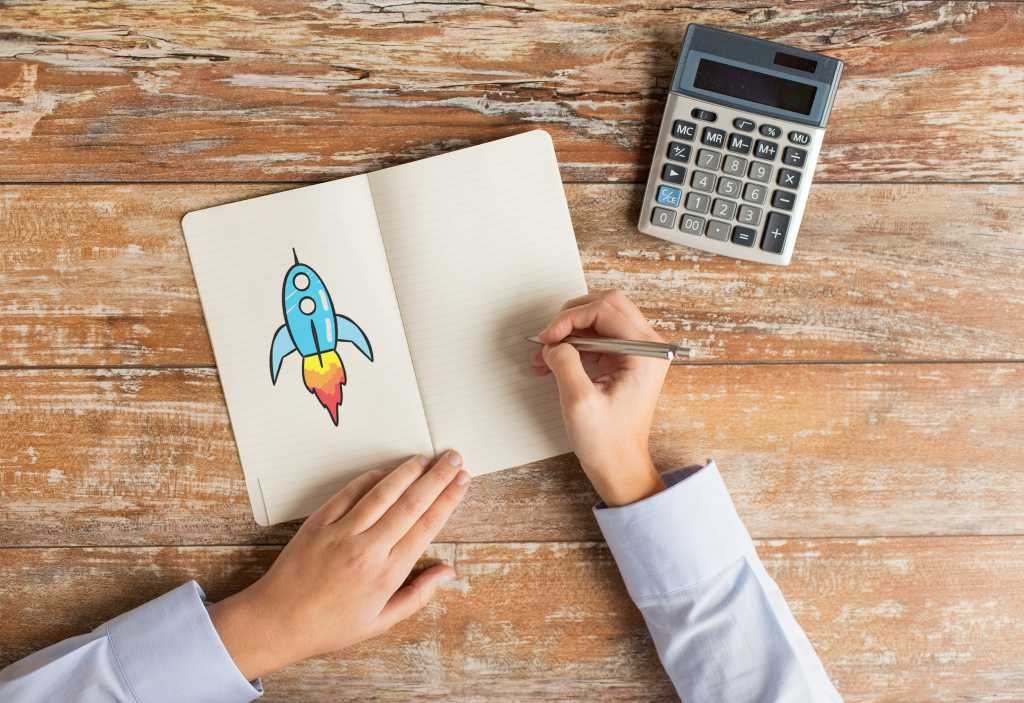 Someone sits at a desk drawing a colorful rocket ship on a paper pad.