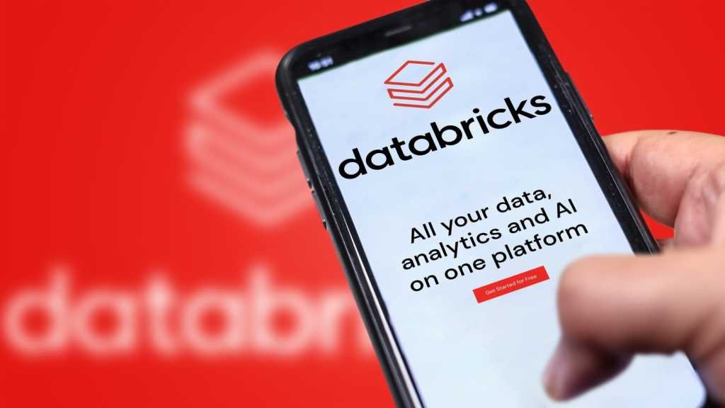 A photograph of a Databricks website on a phone being held in front of a larger, blurry version of the Databricks logo.